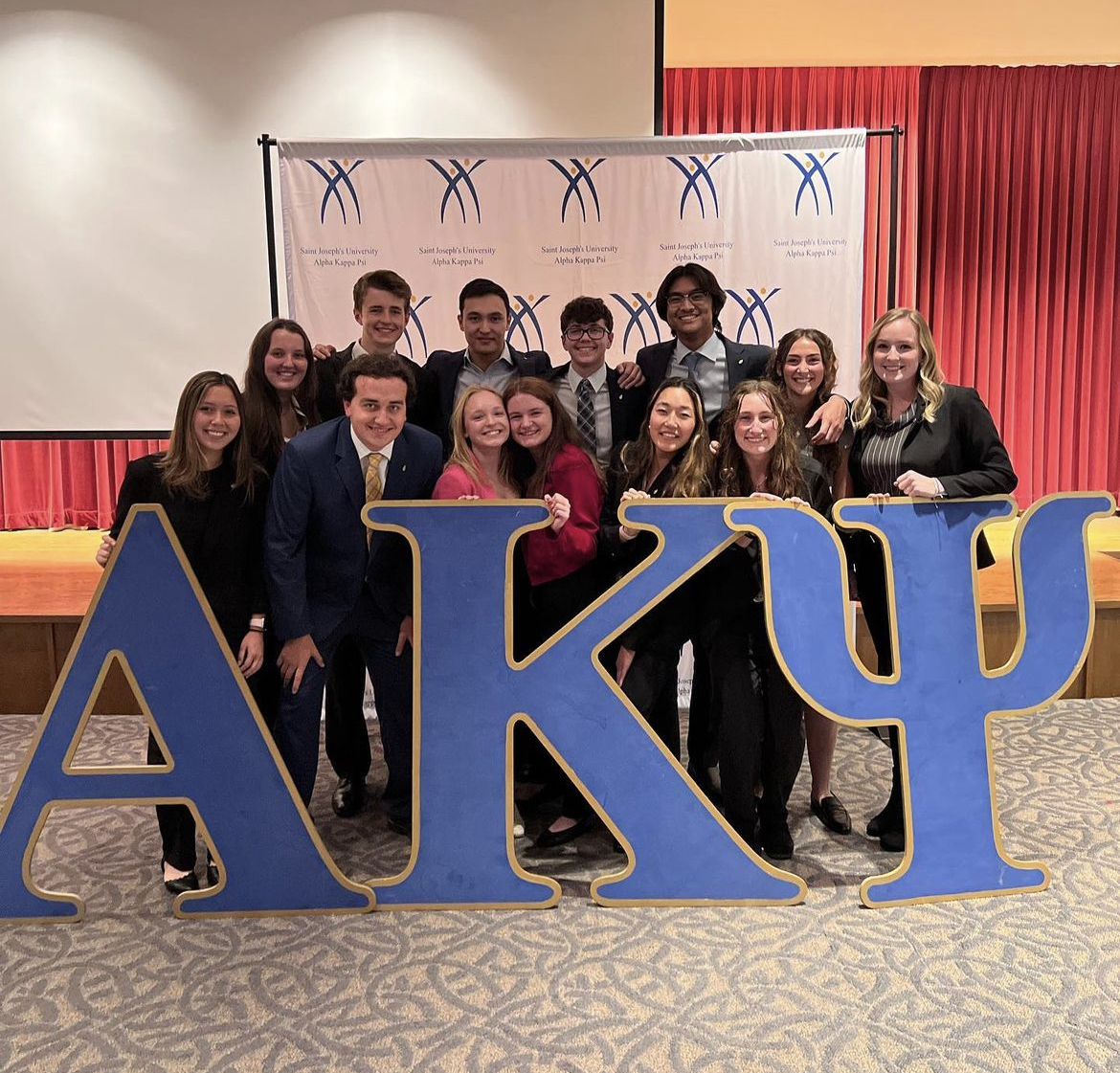 group of 13 college students wearing business prefessional clothing after being inducted into the fraternity alpha kappa psi. They are standing behing 3 large wooden letters a k and p that are blue and yellow. there is a white back drop behind them.